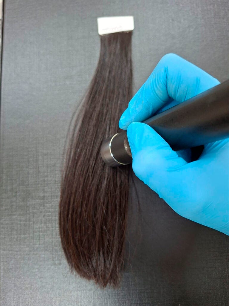 Simulação de mecha de cabelo castanho sendo analisada. Pesquisadora com luvas azuis utiliza o aparelho nas mechas. Simulação de mecha de cabelo castanho sendo analisada. Pesquisadora com luvas azuis utiliza o aparelho nas mechas.