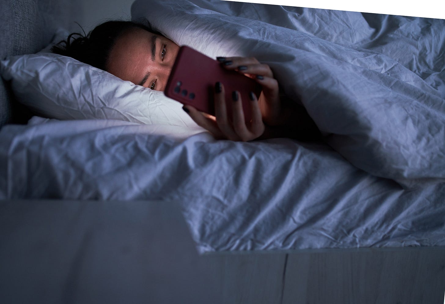 Person in bed in the dark looking at their phone with only the top half of their face visible. Person in bed in the dark looking at their phone with only the top half of their face visible.