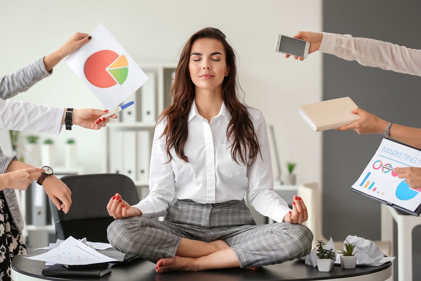 A woman seated on a desk in the lotus pose surrounded by charts and graphs