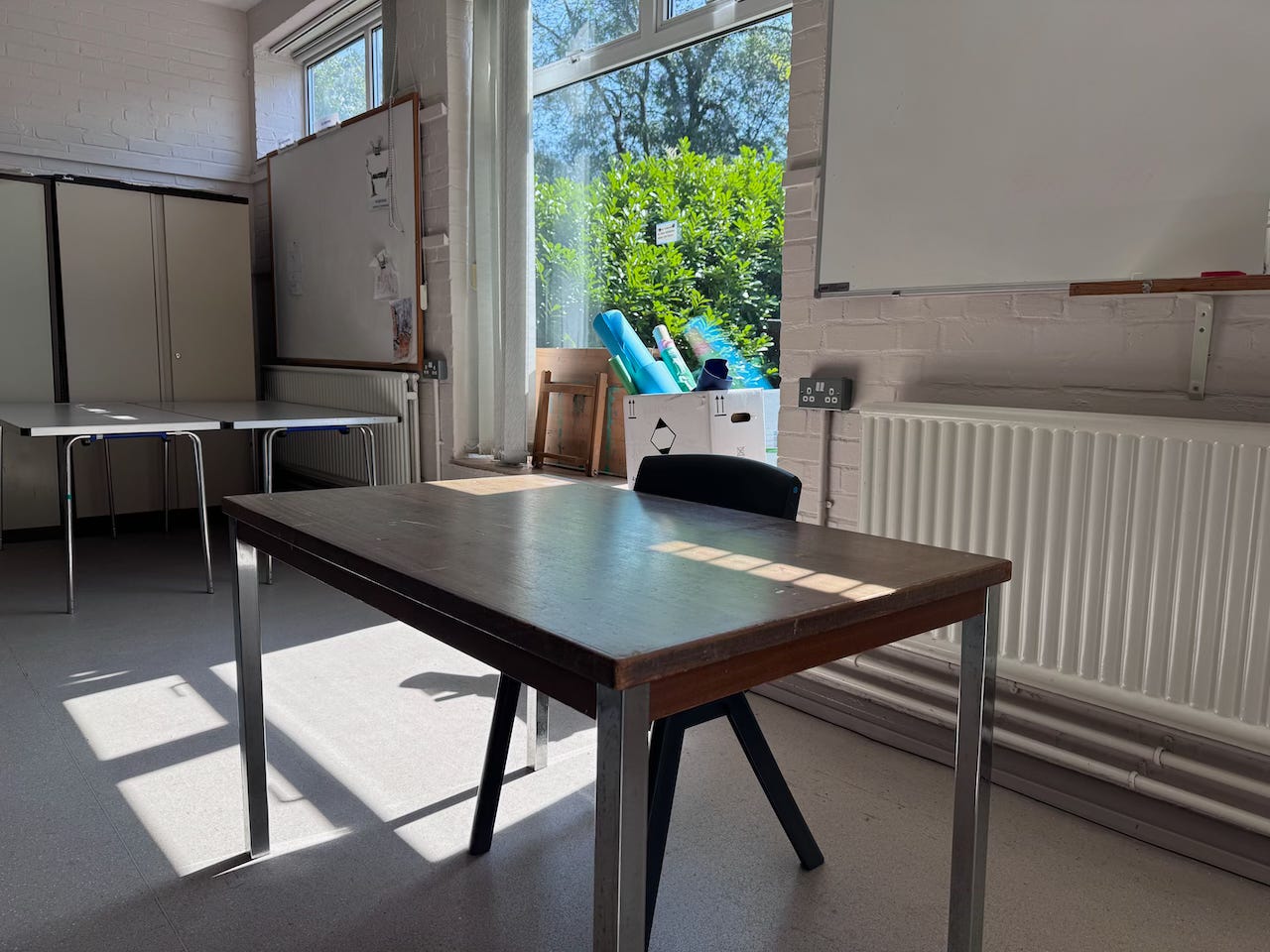 Classroom with large windows. There is a wooden desk and chair just offset from the window. There are squares of light from the sunlight coming through the windows on the table top. 
