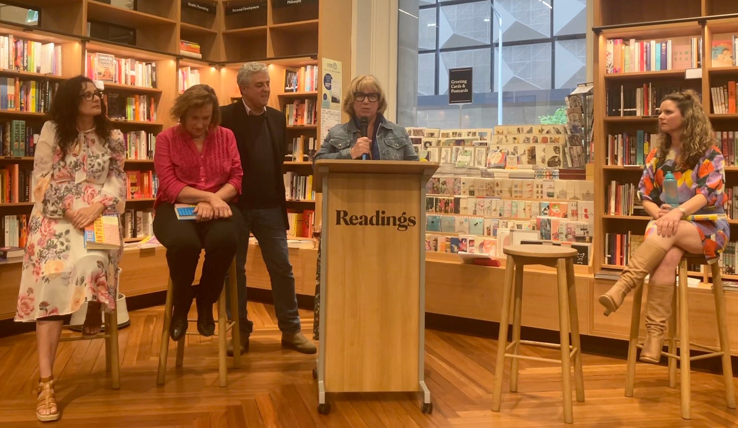 A woman stands at a lectern in a bookstore reading an excerpt from a published story. Three other women authors sit listening after their readings and a man hosting the event watches on.