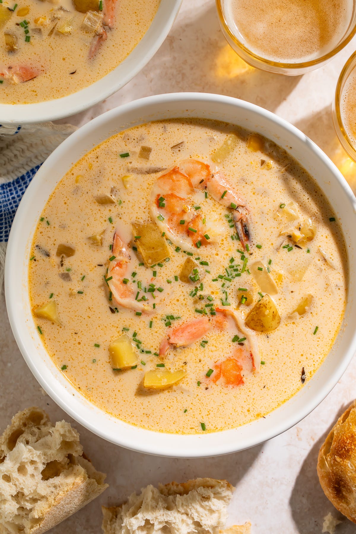 Bowl of creamy chowder with shrimp and potatoes, garnished with snipped chives, surrounded by crusty baguette and glasses of beer