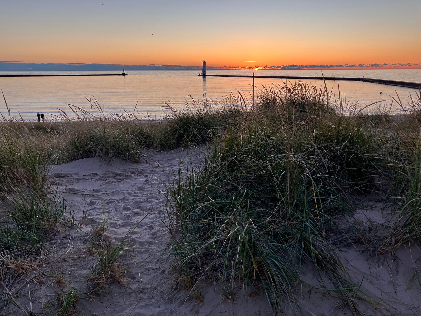A picture of the pier on Lake Michigan, in northern Michigan. Dune grass is in the foreground, with a lighthouse in the distance and a low sunset at the horizon line. A picture of the pier on Lake Michigan, in northern Michigan. Dune grass is in the foreground, with a lighthouse in the distance and a low sunset at the horizon line.