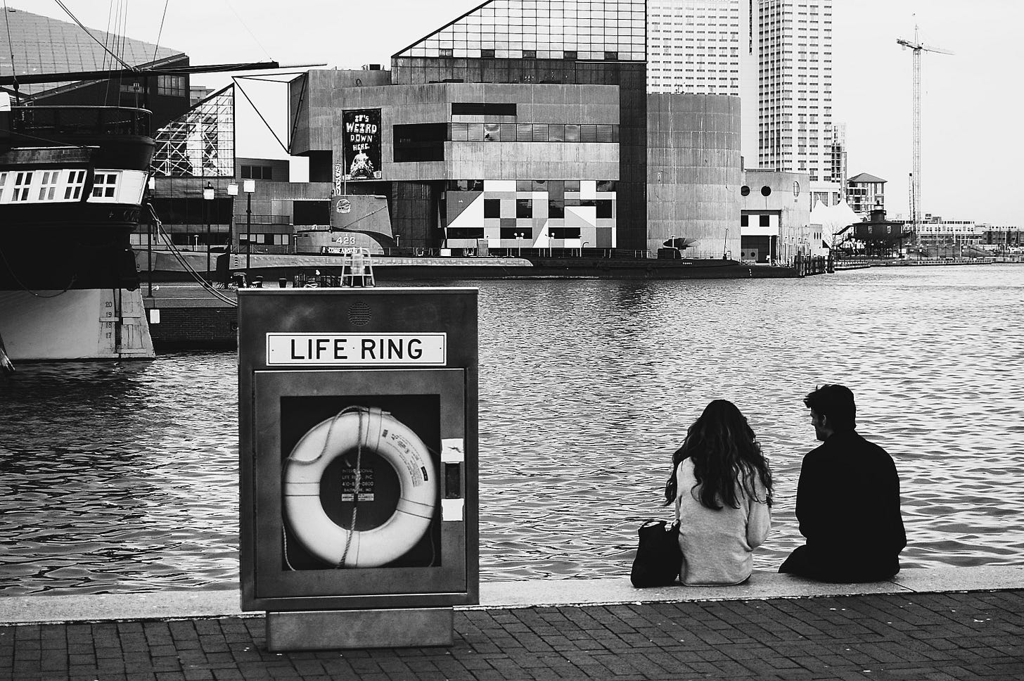 A couple sits on the ledge overlooking the National Aquarium in Baltimore's Inner Harbor. A life preserver sits in a box next to them. A couple sits on the ledge overlooking the National Aquarium in Baltimore's Inner Harbor. A life preserver sits in a box next to them.
