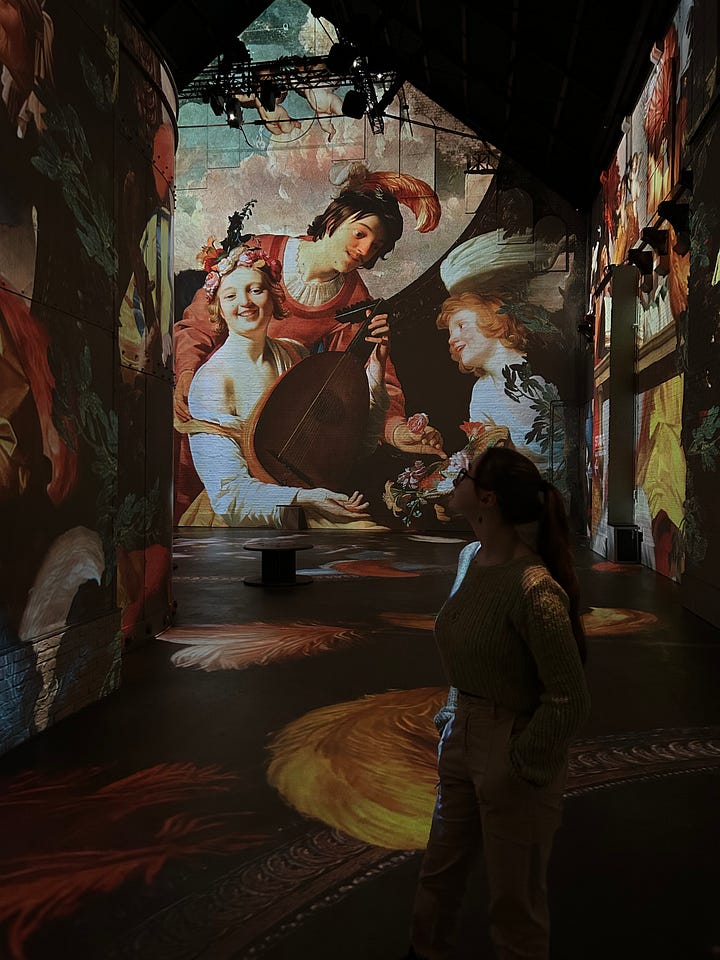 Darker image of a woman walking around an immersive Van Gogh experience, with paintings projected onto walls, and a man walking through a gallery space of art featuring cats