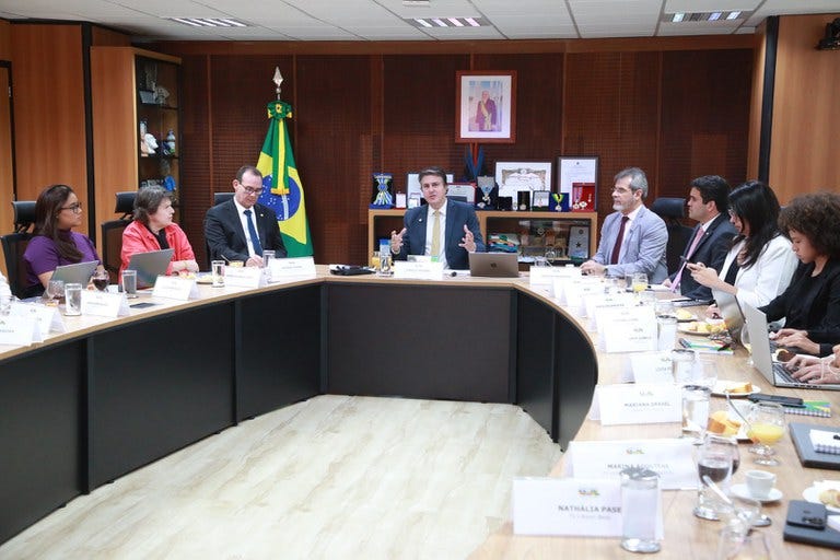 #descrevemospravocê A imagem mostra uma reunião formal em uma sala de governo. Ao redor de uma grande mesa em formato de U, estão sentadas aproximadamente doze pessoas, entre homens e mulheres, em trajes formais. A maioria delas tem frente a si papéis, notebooks ou copos com água e café. A mesa está organizada com placas de identificação de cada participante, além de pratos com frutas e pães no canto direito da imagem, sugerindo um café da manhã de trabalho. No centro da imagem, um homem de terno escuro e gravata amarela fala gesticulando com as mãos. Atrás dele, vê-se uma parede de madeira, com prateleiras que exibem livros, troféus e objetos decorativos. À sua esquerda está posicionada uma bandeira do Brasil, e acima dela, um quadro com uma fotografia oficial. A sala tem iluminação fluorescente e teto rebaixado com placas brancas. O ambiente transmite formalidade institucional, típico de reuniões ministeriais ou encontros de gestores públicos em espaços governamentais. #descrevemospravocê A imagem mostra uma reunião formal em uma sala de governo. Ao redor de uma grande mesa em formato de U, estão sentadas aproximadamente doze pessoas, entre homens e mulheres, em trajes formais. A maioria delas tem frente a si papéis, notebooks ou copos com água e café. A mesa está organizada com placas de identificação de cada participante, além de pratos com frutas e pães no canto direito da imagem, sugerindo um café da manhã de trabalho. No centro da imagem, um homem de terno escuro e gravata amarela fala gesticulando com as mãos. Atrás dele, vê-se uma parede de madeira, com prateleiras que exibem livros, troféus e objetos decorativos. À sua esquerda está posicionada uma bandeira do Brasil, e acima dela, um quadro com uma fotografia oficial. A sala tem iluminação fluorescente e teto rebaixado com placas brancas. O ambiente transmite formalidade institucional, típico de reuniões ministeriais ou encontros de gestores públicos em espaços governamentais.