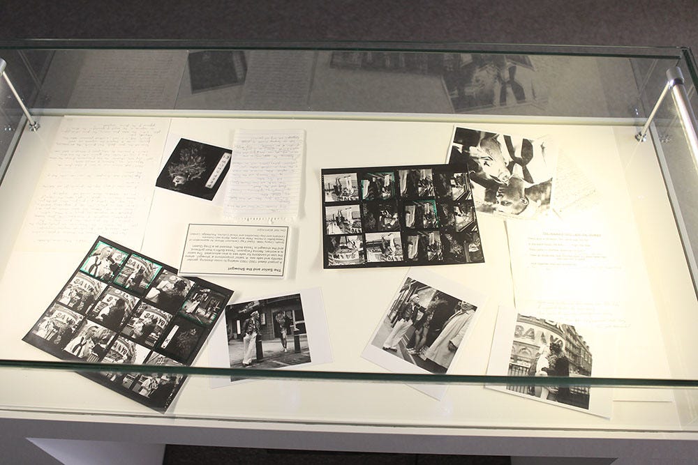 A glass display case contains black and white photographs and handwritten notes. The arrangement suggests a historical or artistic exhibit, evoking nostalgia. There is a text card in the center with context about Tessa's photos, notes, and inspirations.