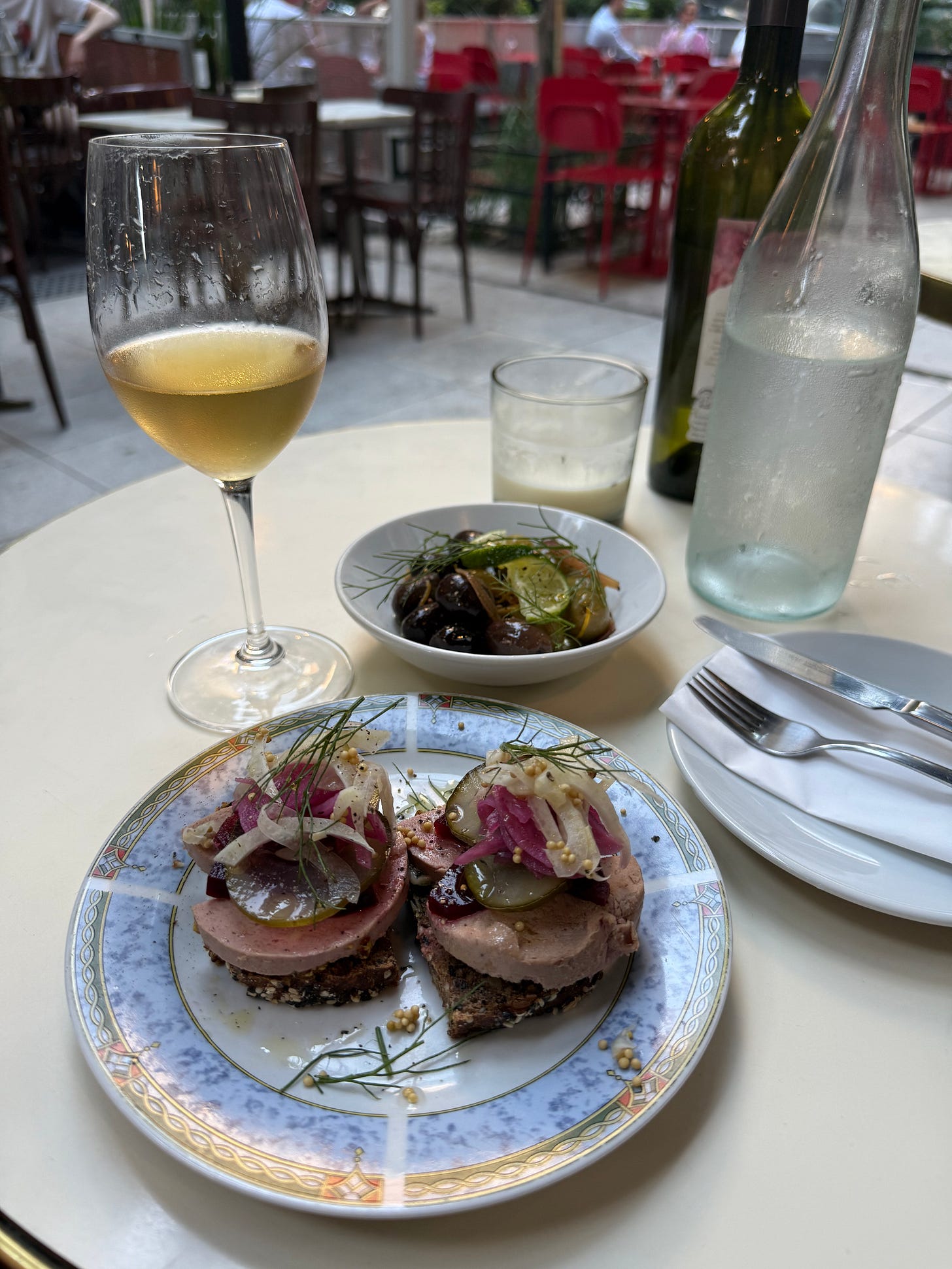 Table with a glass of white wine, a German open faced sandwich of liverwurst and pickled vegetables and a small dish of olives.. and a large bottle of cold water. at a cafe with outside eating in Buenos Aires.