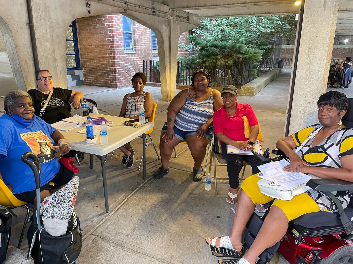 Six Black women sit around a card table under a concrete portico, with a brick building and foliage behind them. Six Black women sit around a card table under a concrete portico, with a brick building and foliage behind them.
