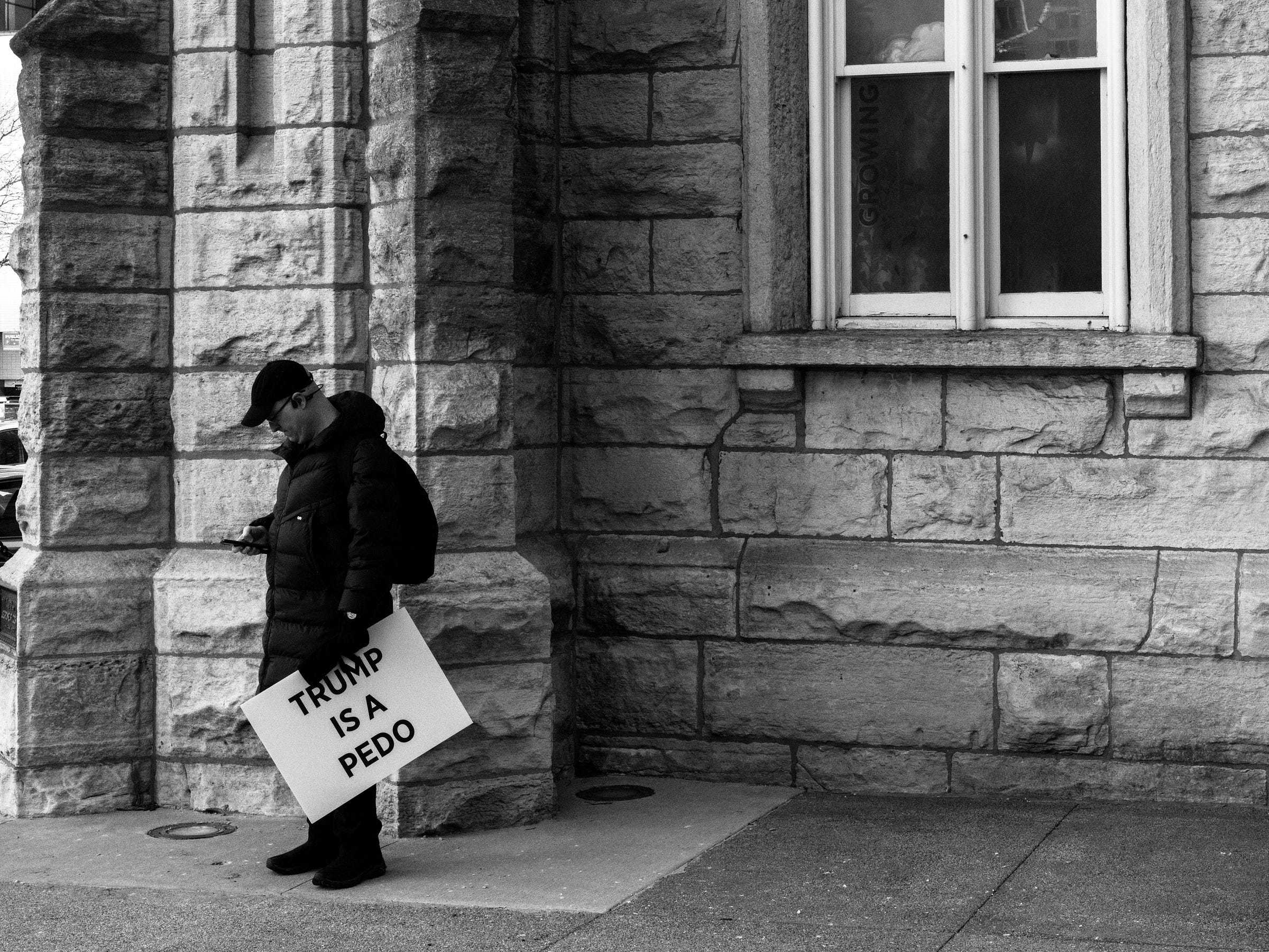 A black and white photograph captures a solitary protester leaning against the stone wall of a historic building. Dressed in a black puffer jacket, hat, and gloves, they are engrossed in their phone while holding a protest sign that reads, "TRUMP IS A PEDO." The stark contrast of the stone architecture and the lone figure creates a contemplative, almost quiet moment amid the larger protest. A window above reflects faint interior details, adding depth to the otherwise still scene. A black and white photograph captures a solitary protester leaning against the stone wall of a historic building. Dressed in a black puffer jacket, hat, and gloves, they are engrossed in their phone while holding a protest sign that reads, "TRUMP IS A PEDO." The stark contrast of the stone architecture and the lone figure creates a contemplative, almost quiet moment amid the larger protest. A window above reflects faint interior details, adding depth to the otherwise still scene.