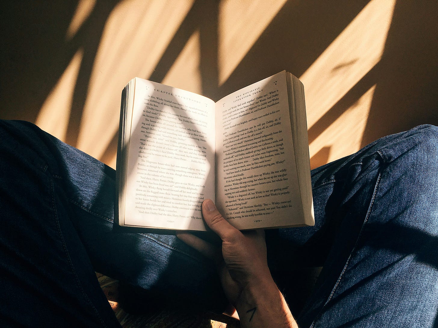 A person reads an open book while sitting down cross-legged. They are wearing blue jeans. The floor is brown wood with shadows and sunlight coming in through a nearby window.