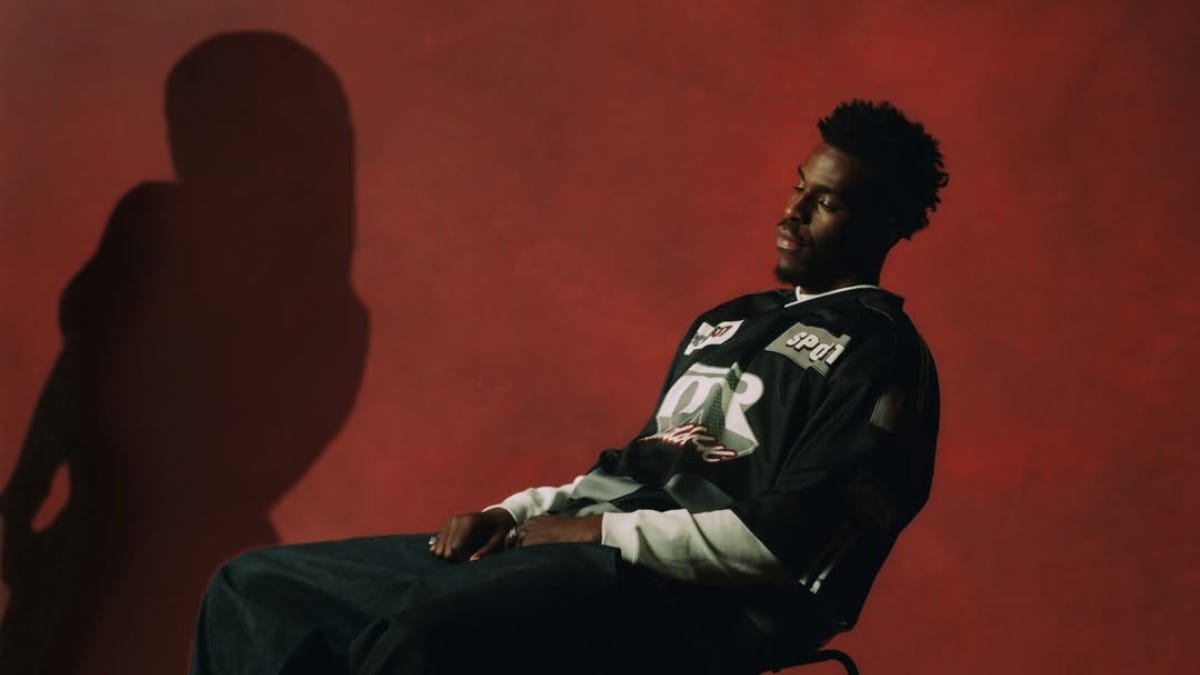 A man sits reclined in a chair against a deep red background, lit dramatically from one side. He wears a dark graphic jersey layered over a long-sleeve shirt and jeans. A shadowy silhouette of another figure appears on the wall beside him, adding a moody, cinematic feel. The overall composition feels contemplative and introspective.