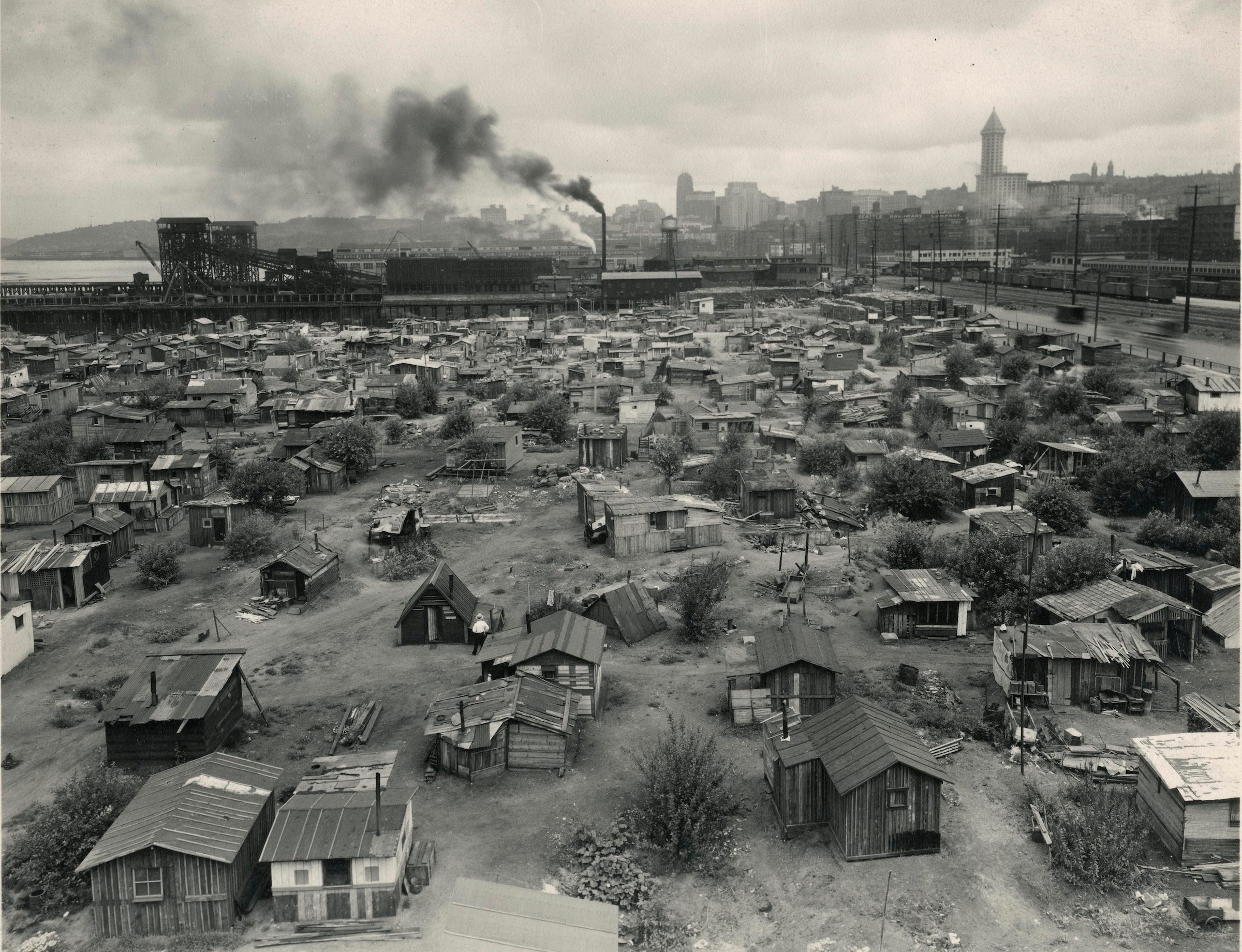 Low angle aerial view of shack village with industrial smoke stacks billowing on the horizon.