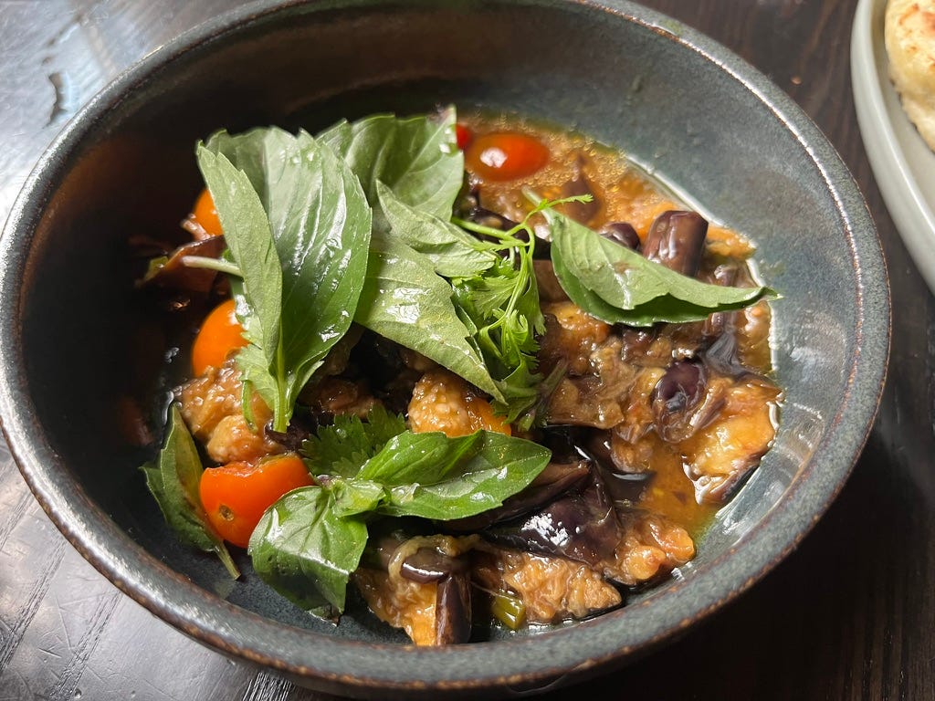 pieces of eggplant with purple skin and yellow flesh and a few whole cherry tomatoes in a yellowish liquid, with many basil leaves on top, all in a gray bowl