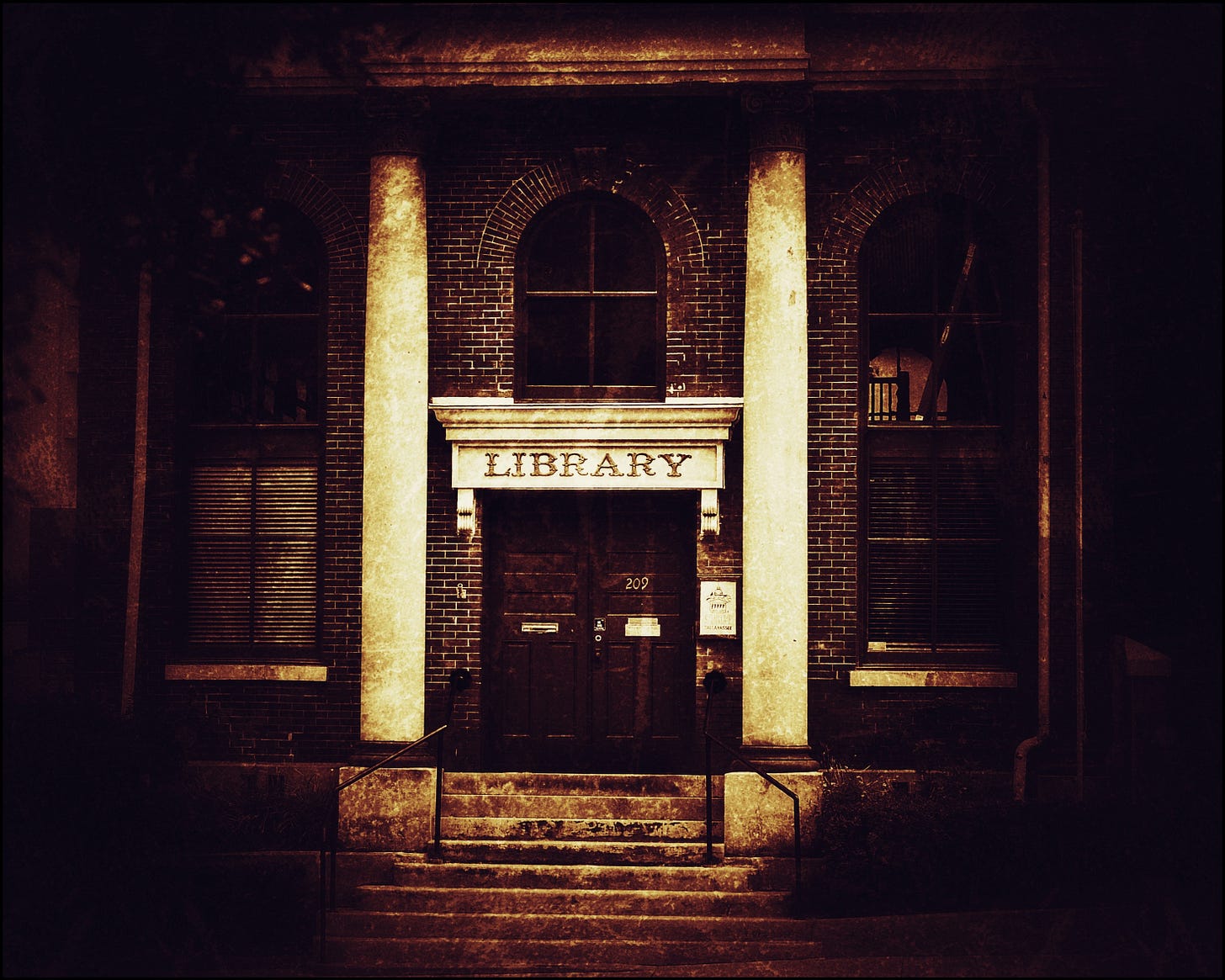 A dark, murky, sepia-tinted photo of the front of an old brick library. There's a short flight of concrete stairs leading up to the entrance; to either side is a substantial stone column. Over the entrance door a single word is carved: "LIBRARY."