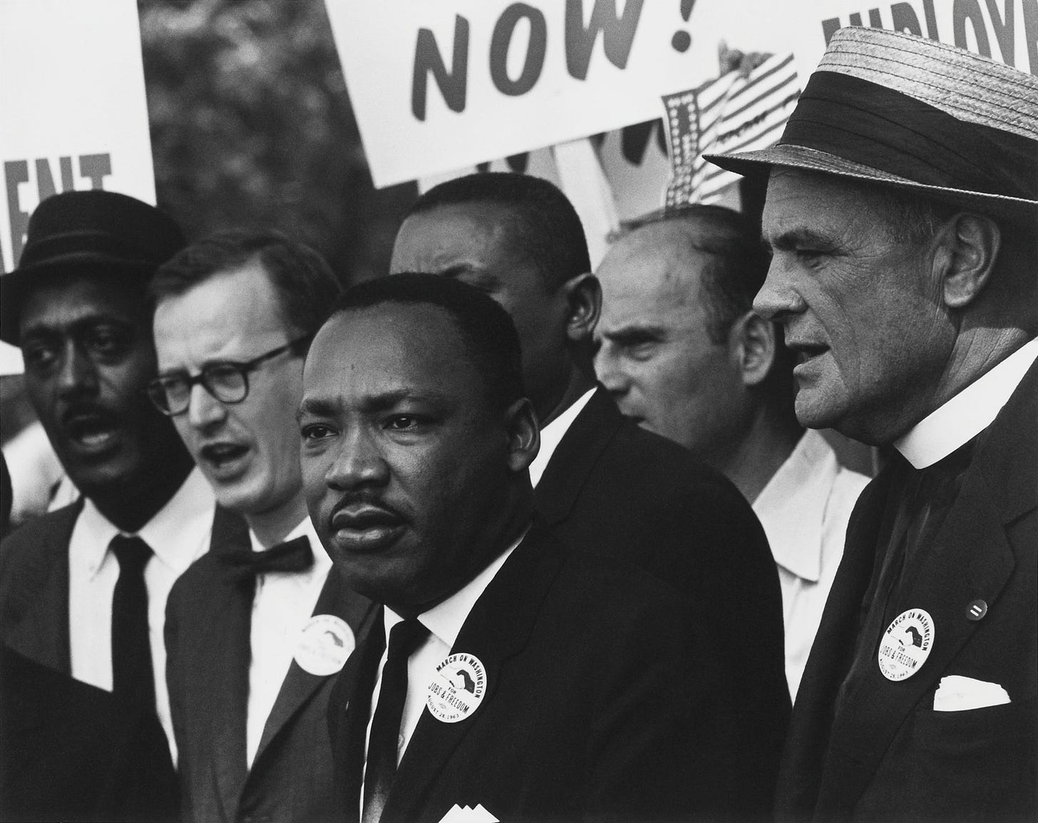 A black and white photo from 1963 showing Dr. Martin Luther King Jr. standing in a tight crowd at the March on Washington. He is wearing a dark suit and looking off to the side with a serious expression. He is surrounded by other activists in suits and hats, including Mathew Ahmann. In the background, protest signs are partially visible.