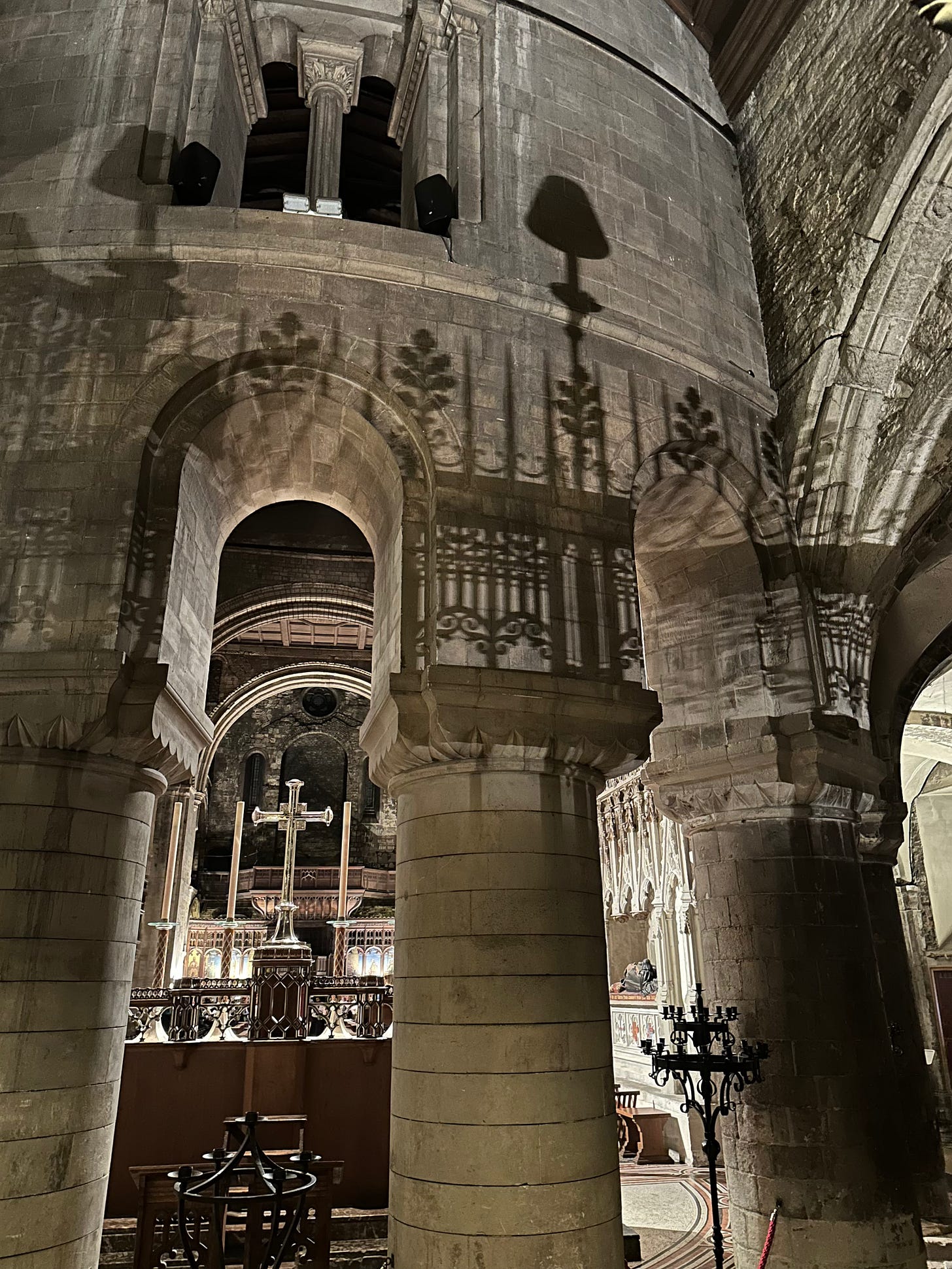 Stone arches and thick columns inside a historic church, with patterned shadows cast on the walls and a lit altar and cross visible beyond.