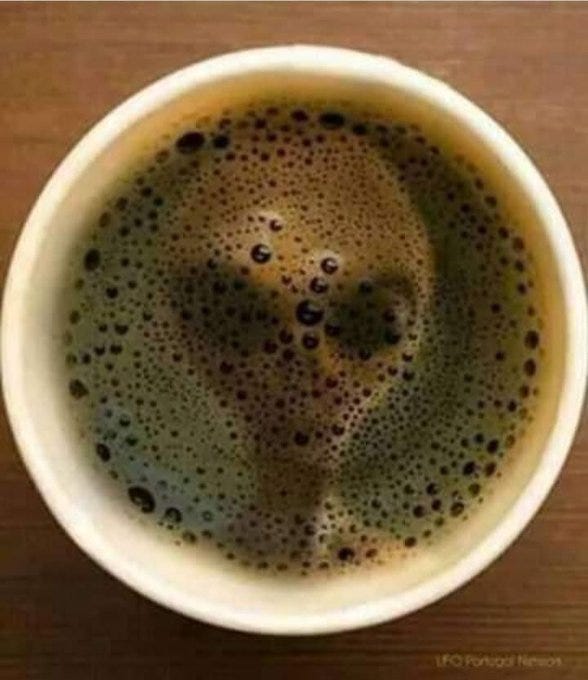 A white ceramic cup filled with dark brown coffee sits on a wooden table. The surface of the coffee features bubbly foam that naturally forms a humanoid face shape with two dark eye-like spots and a mouth-like curve below, creating a pareidolia effect. The foam bubbles are clustered to outline the facial features clearly.
