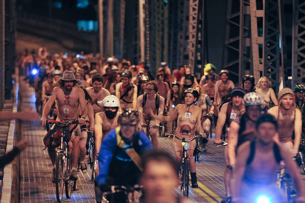 A large group of mostly nude cyclists ride together across Portland’s Hawthorne Bridge at night, many wearing helmets and decorated with body paint.