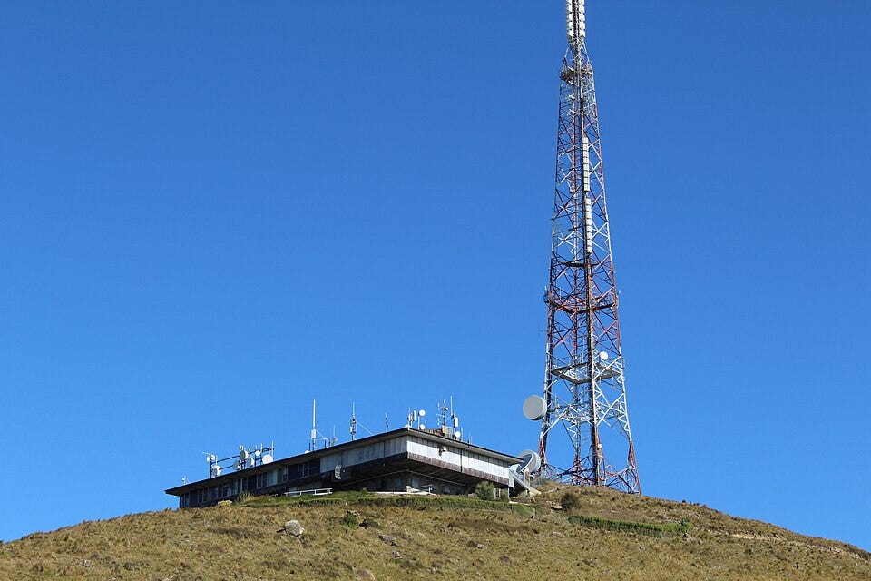 A telecommunications and broadcast facility sits on a dry grassy hill beneath a tall red-and-white transmission tower. The low industrial building is covered with antennas and satellite dishes, with a clear blue sky filling the background.