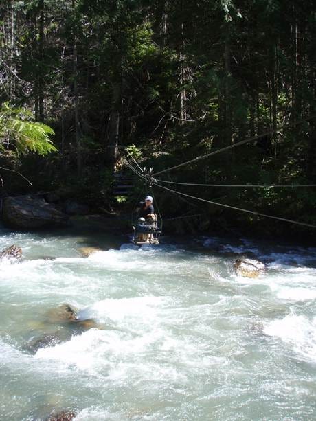 A woman and a dog sit inside an open metal cable car crossing a rushing creek. The bank behind her is dark with trees, and the waters rush white beneath her.