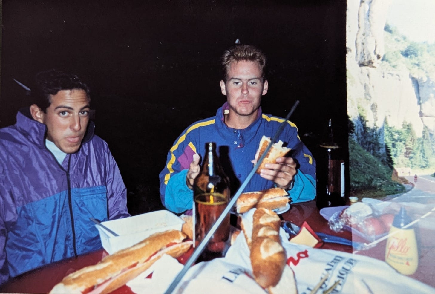 Two young men are standing near the roof of a red rental car. On the roof of the car are baguettes, with ham and cheese and mustard and tomatoes and beer. The men are wearing windbreakers and it looks cold.