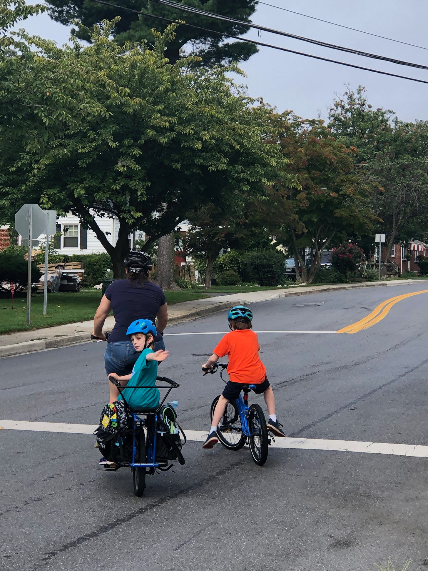 Me (a white woman in a navy shirt) on my cargo bike with my younger son (a white boy in a teal shirt) on the back waving and my older son (a white boy in an orange shirt and navy shorts) on his own bike