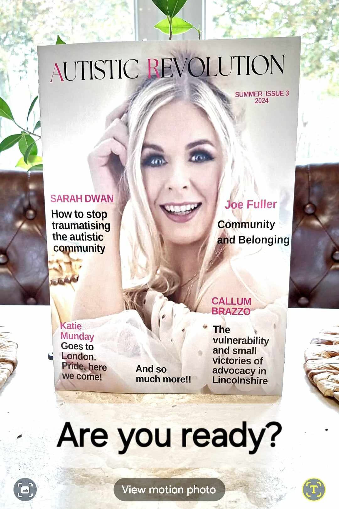 Photo of a printed copy of the Autistic Revolution magazine! The magazine is in an upright position on a counter and in front of a window. The main image on the cover is of Sarah Dwan who is a pale person, with long blonde hair and is smiling widely at the viewer.