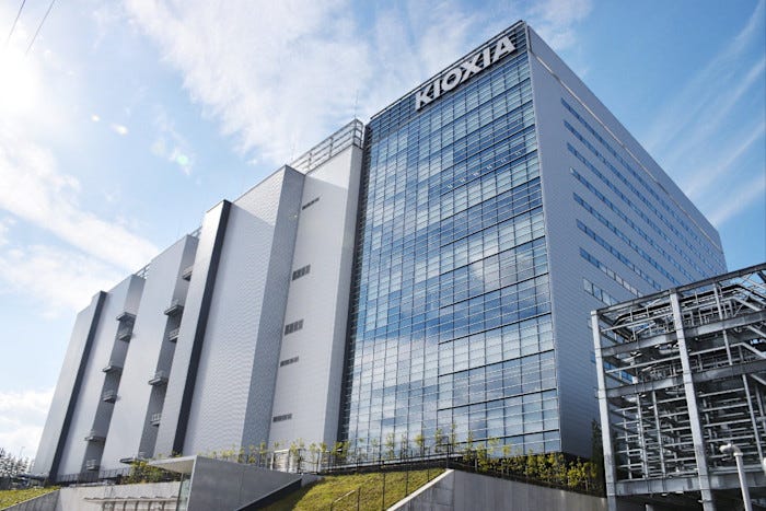 The Kitakami factory of Kioxia, a large modern building with glass and metal exterior, under a partly cloudy sky.