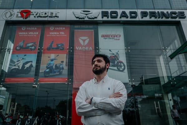 Portrait of Muhammad Salman standing with arms crossed in front of a dealership decorated with posters of motorcycles and scooters for sale.