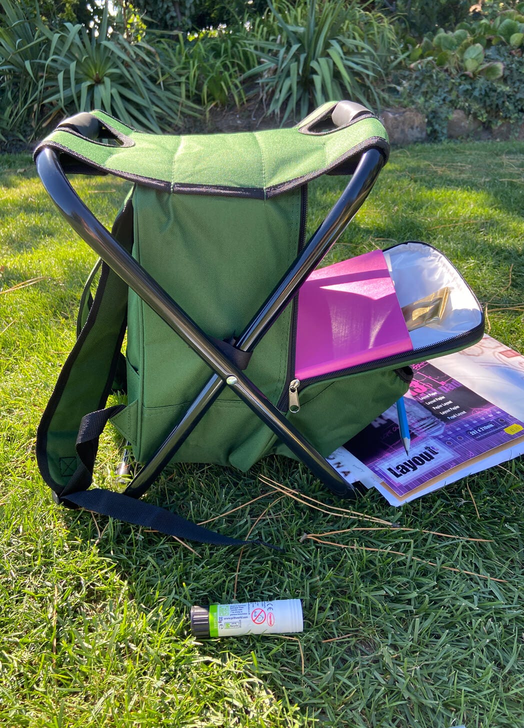 photo of a backpack that doubles as a stool. Art materials are visible in the bags pocket. It's standing on grass in a public park