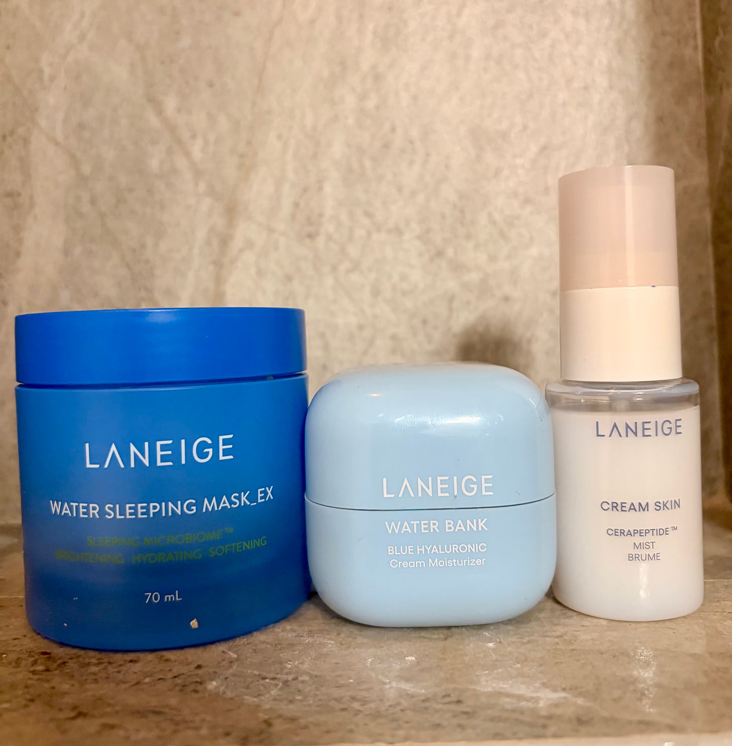 A medium shot photograph of three distinct Laneige skincare products standing on a light marble surface. From left to right: a large, deep blue tub of Water Sleeping Mask_EX (70 mL), a smaller light blue, squared-off jar of Water Bank Blue Hyaluronic Cream Moisturizer, and a sleek, tall white pump bottle of Cream Skin Cerapeptide Mist. The Laneige logo and product functions (hydrating, softening) are clearly visible on the packaging. A medium shot photograph of three distinct Laneige skincare products standing on a light marble surface. From left to right: a large, deep blue tub of Water Sleeping Mask_EX (70 mL), a smaller light blue, squared-off jar of Water Bank Blue Hyaluronic Cream Moisturizer, and a sleek, tall white pump bottle of Cream Skin Cerapeptide Mist. The Laneige logo and product functions (hydrating, softening) are clearly visible on the packaging.