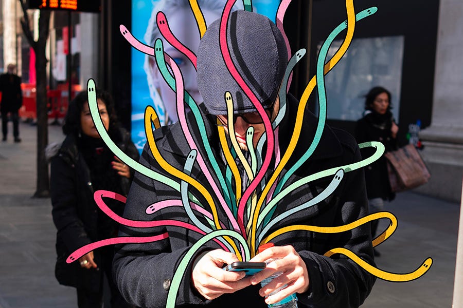 Photograph of a man looking down at his smartphone while colorful, surreal illustrated shapes and characters spiral out of the device and encircle his head, symbolizing the competing demands of digital attention.