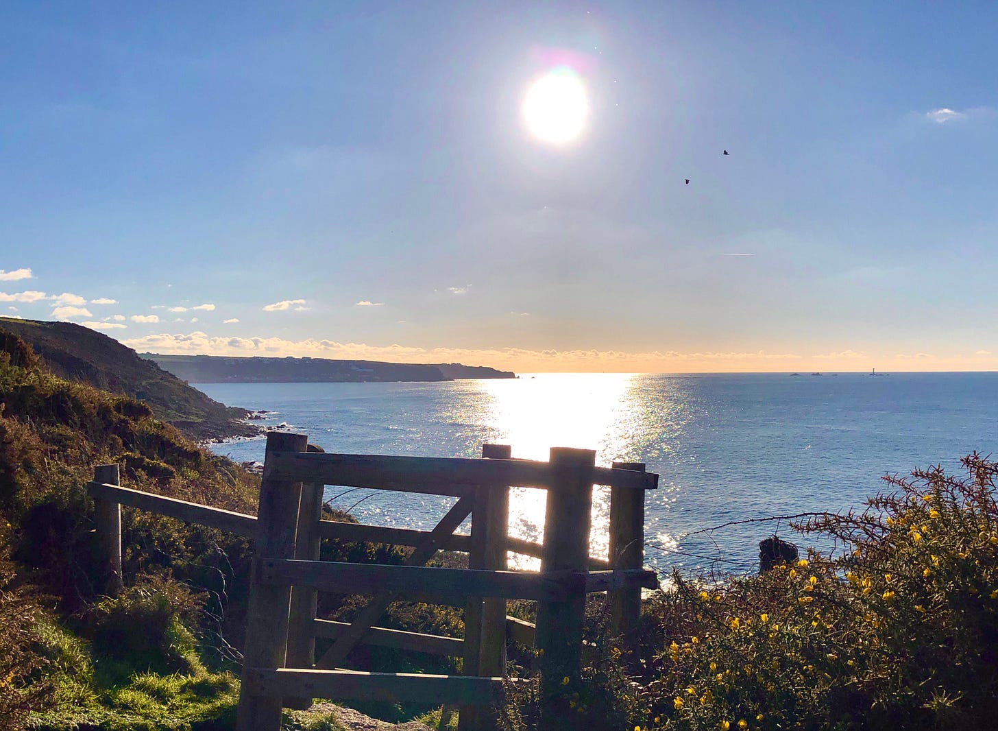 A wooden gate on the coast path. The sun is shining brightly and reflecting off the blue sea.