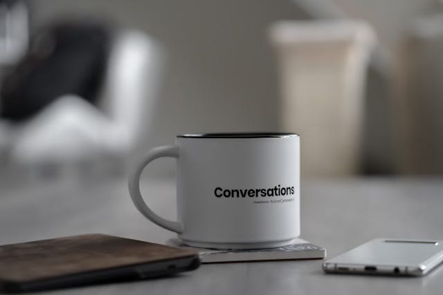 A white mug with the word Conversations on the side, resting on a coaster on a white desk.