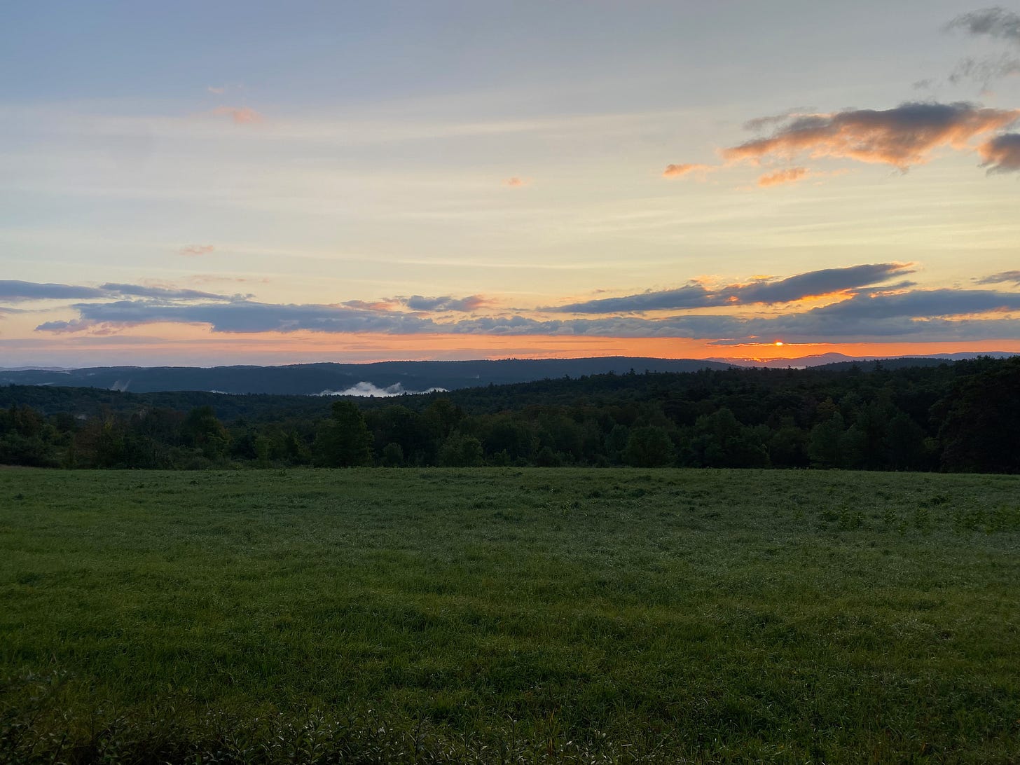 Sunrise over a grassy ridgetop pasture. Wisps of clouds sit in the mountains and the sun is a tiny golden orb among purple and orange clouds.