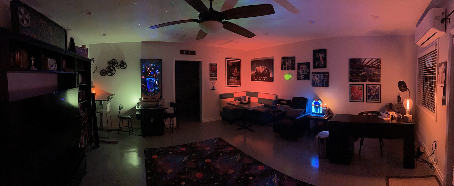 additional dwelling unit turned into a writers office, with a desk and chair to the right, an easy chair, small table, and mini jukebox in the center, a diner booth in the left corner. there is artwork on the walls and an arcade rug with cartoon planets, a ceiling fan overhead. the whole thing is lit by a few small colored lights, orange, green, and blue.