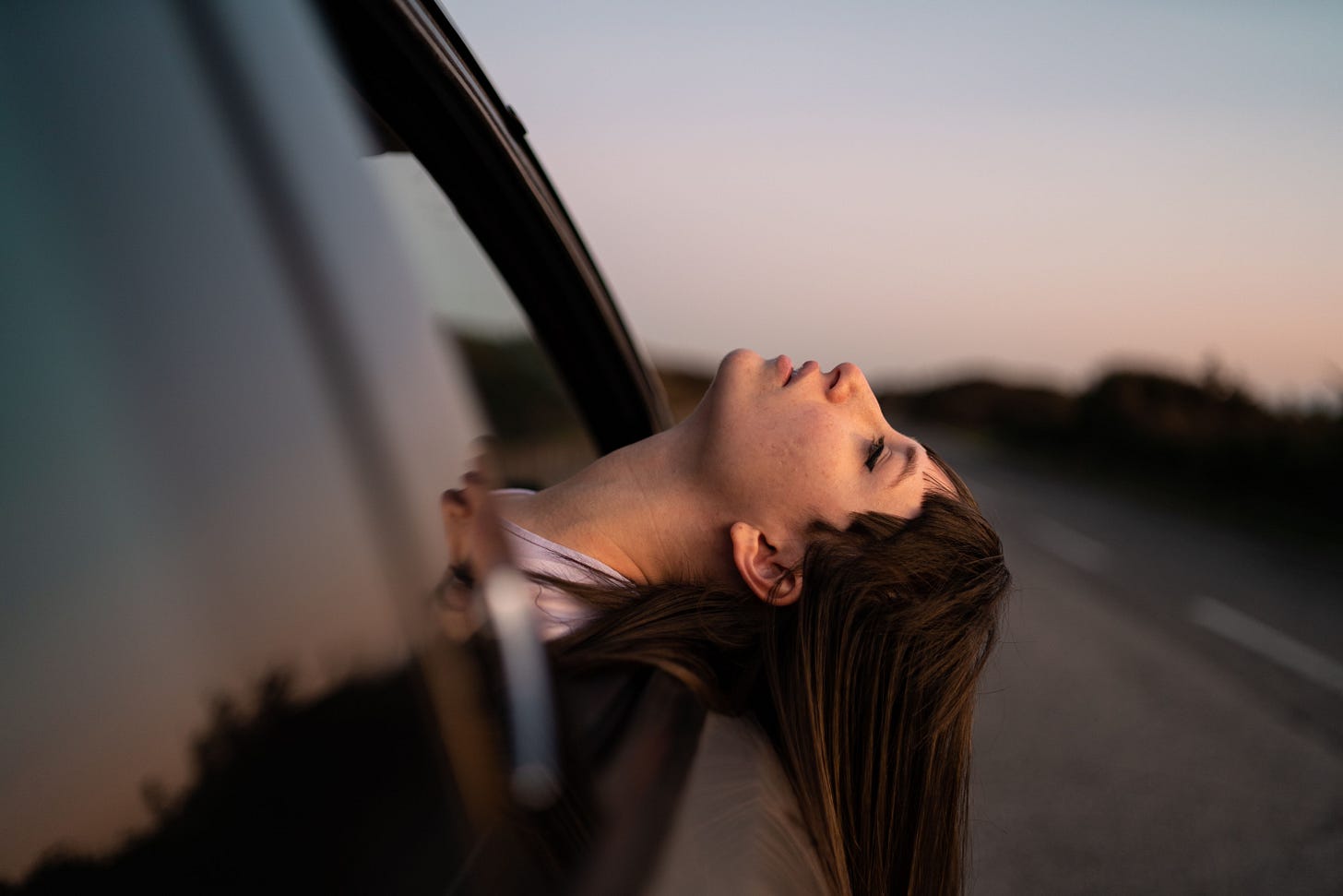 Eine Frau sitzt in einem Auto und posiert mit dem Kopf aus dem offenen Fenster. Die Augen sind geschlossen und der Blick nach oben gerichtet. Sie hat braune, lange Haare, die an der Seite der Autotür entlang Richtung Boden fließen.