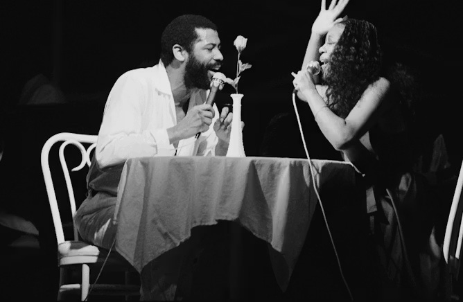 Black-and-white photo of couple singing while seated at a round dining table with a rose in a vase in the center