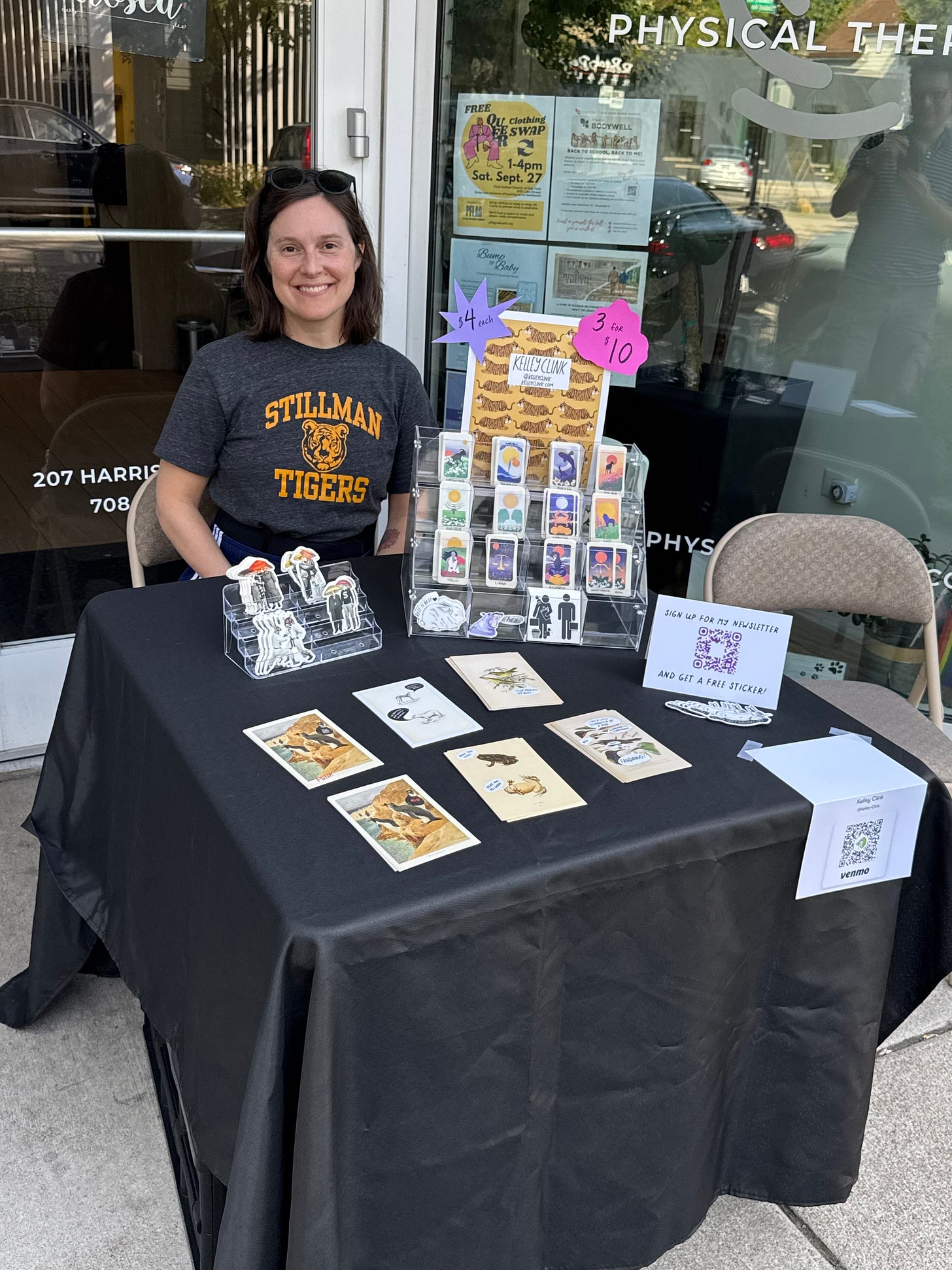 Kelley sitting at their table with all of their stickers displayed. They have sunglasses on their head, and are smiling, but honestly they look pretty tired. 