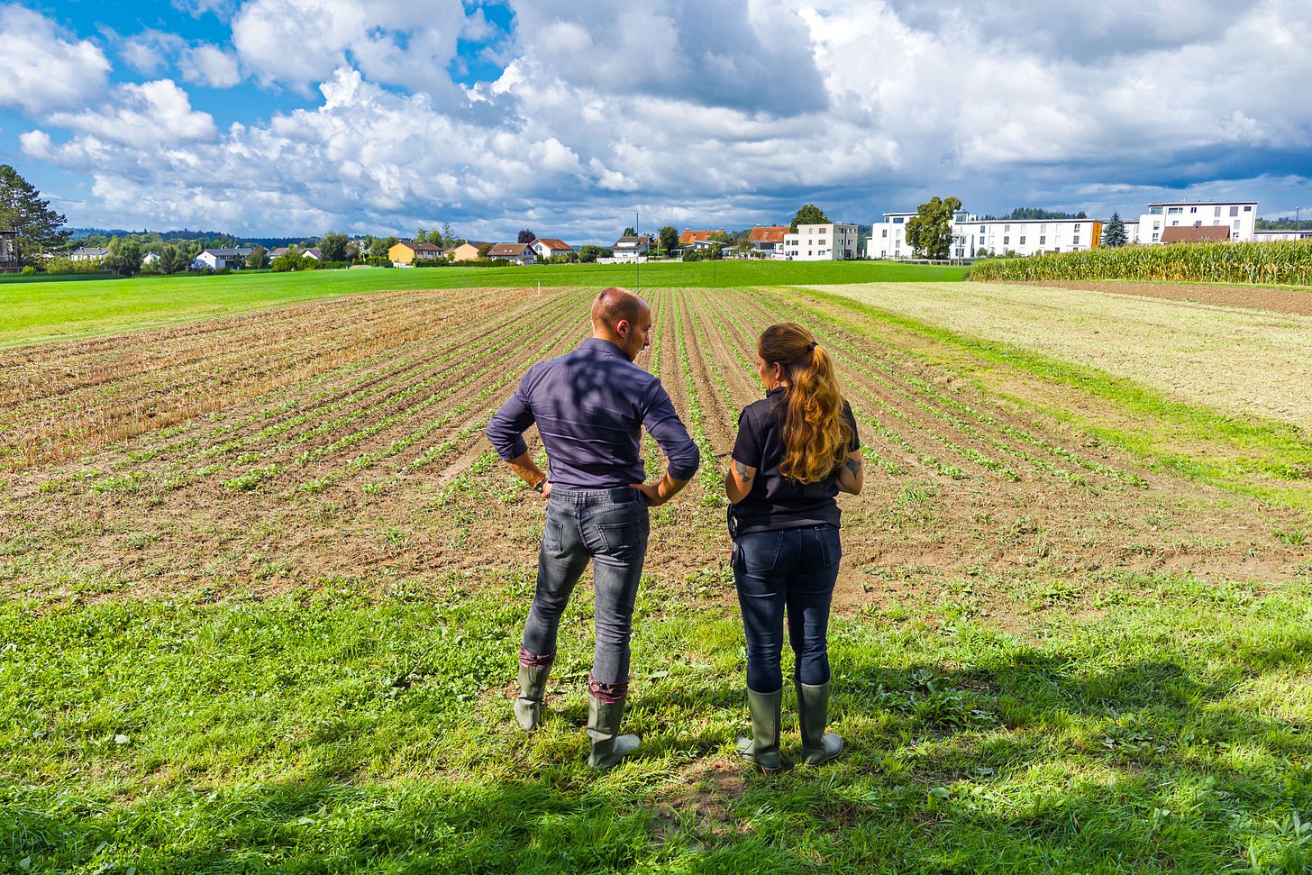 Zwei Studierende in Gummistiefeln stehen auf dem Oberacker in Zollikofen und blicken über frisch bestellte Reihen eines Versuchsfeldes. Im Hintergrund sind weitere Felder, Maiskulturen sowie Wohngebäude am Ortsrand zu sehen, darüber ein Himmel mit imposanten Wolkenformationen.