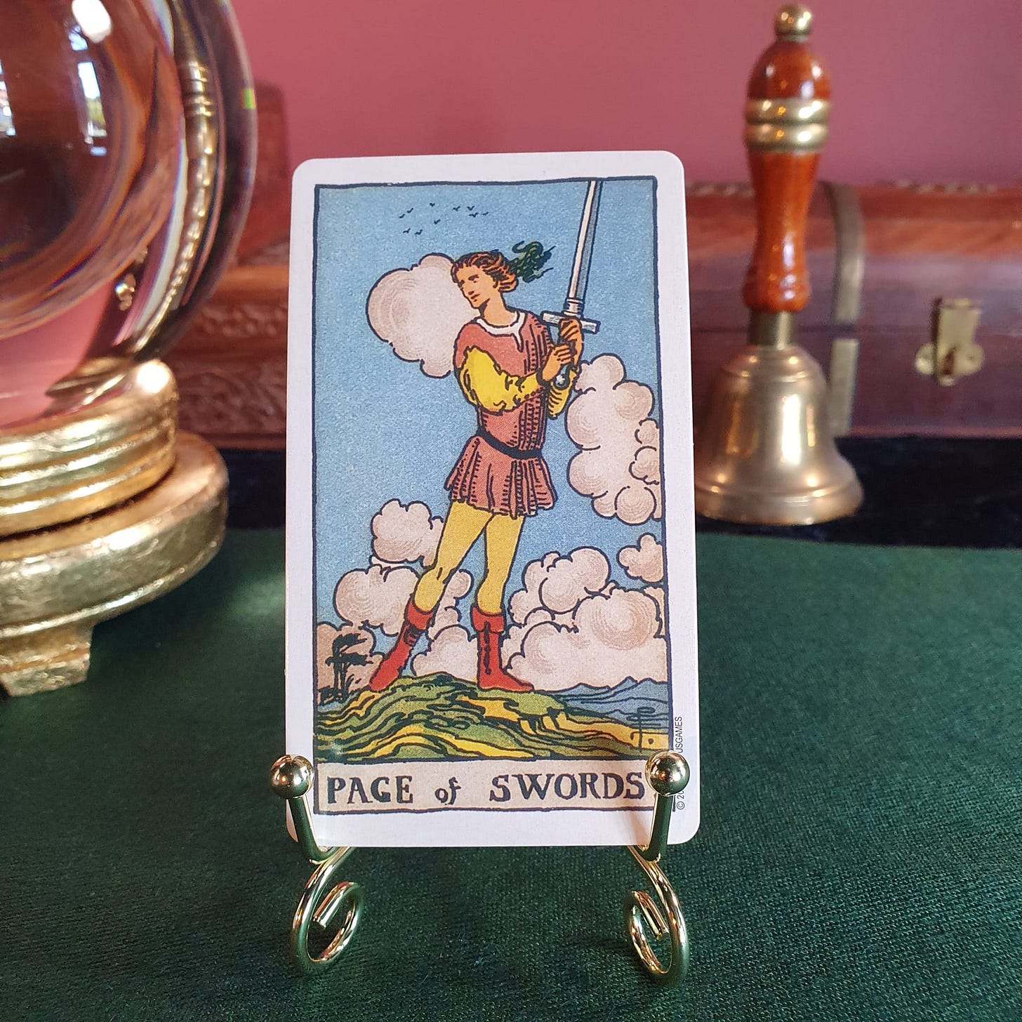 The Page of Swords tarot card from the Smith-Waite deck displayed on a small gold stand, showing a figure holding a sword among clouds and windblown hills.