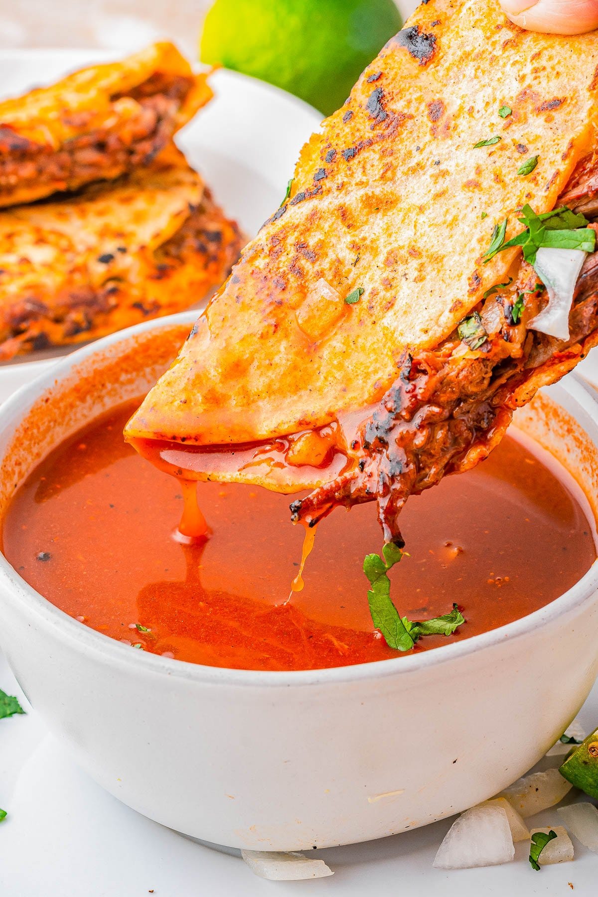 A quesataco filled with shredded meat is being dipped into a bowl of red consommé, with chopped onions and cilantro visible on the taco and plate.