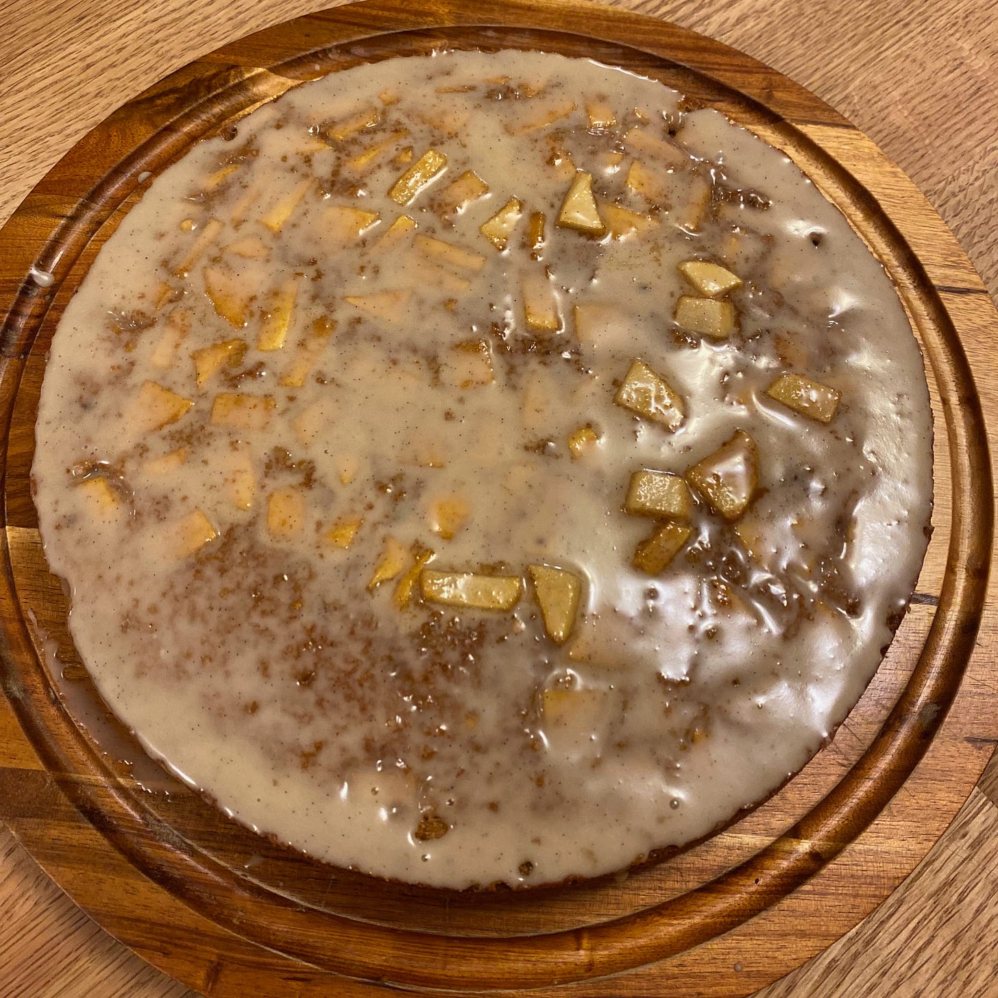 A round cake covered in a shiny white glaze and studded with chunks of apple, sits on a wooden cake plate. 