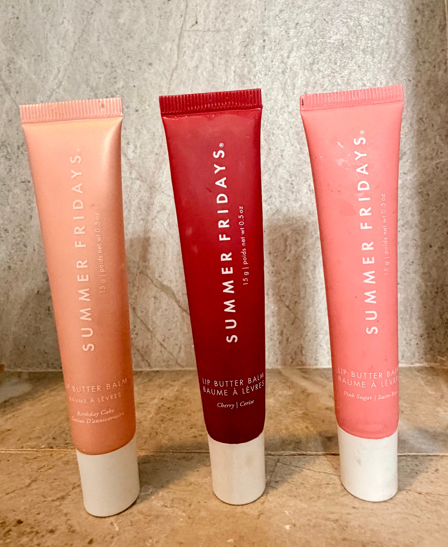 A close-up, medium-shot photograph of three tubes of Summer Fridays Lip Butter Balm standing upright on a textured, light gray concrete-like surface. From left to right: the nude-peach 'Birthday Cake' (marked gâteau d'anniversaire), the deep berry 'Cherry' (marked cerise), and the classic light pink 'Pink Sugar' (marked Sucre Rose). Each tube is 15 g / 0.5 oz with a white twist-off base. The background is a soft, blurred concrete texture. A close-up, medium-shot photograph of three tubes of Summer Fridays Lip Butter Balm standing upright on a textured, light gray concrete-like surface. From left to right: the nude-peach 'Birthday Cake' (marked gâteau d'anniversaire), the deep berry 'Cherry' (marked cerise), and the classic light pink 'Pink Sugar' (marked Sucre Rose). Each tube is 15 g / 0.5 oz with a white twist-off base. The background is a soft, blurred concrete texture.
