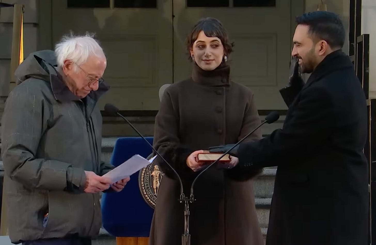 Sanders (elderly white man in puffy coat) reading from paper on left; Duwalji, (young Middle Eastern woman in stylish coat), looking amused and holding book; Mamdani, (young bearded Middle Eastern man in black coat) has hand on book and other hand raised  