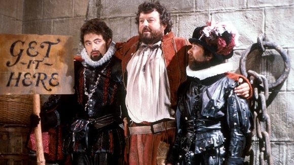 Rowan Atkinson, carrying a sign saying ‘Get it here’, a large bearded sailor who misses his mummy, and Baldrick in Black Adder II, Money