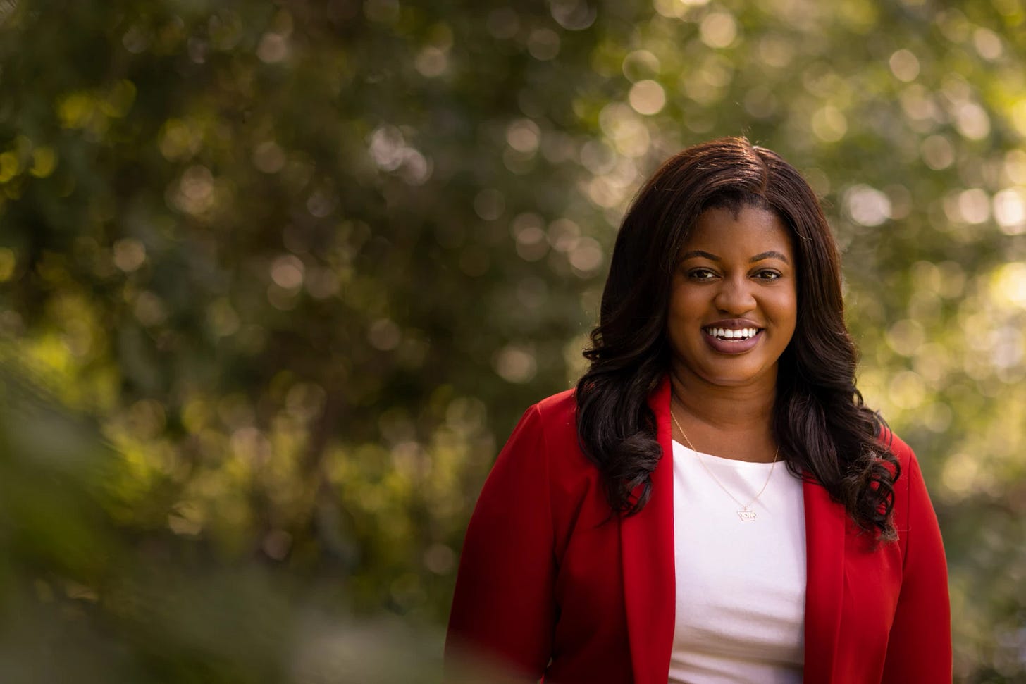 Photo of Deidre DeJear in a red blazer with muted green trees in the background 