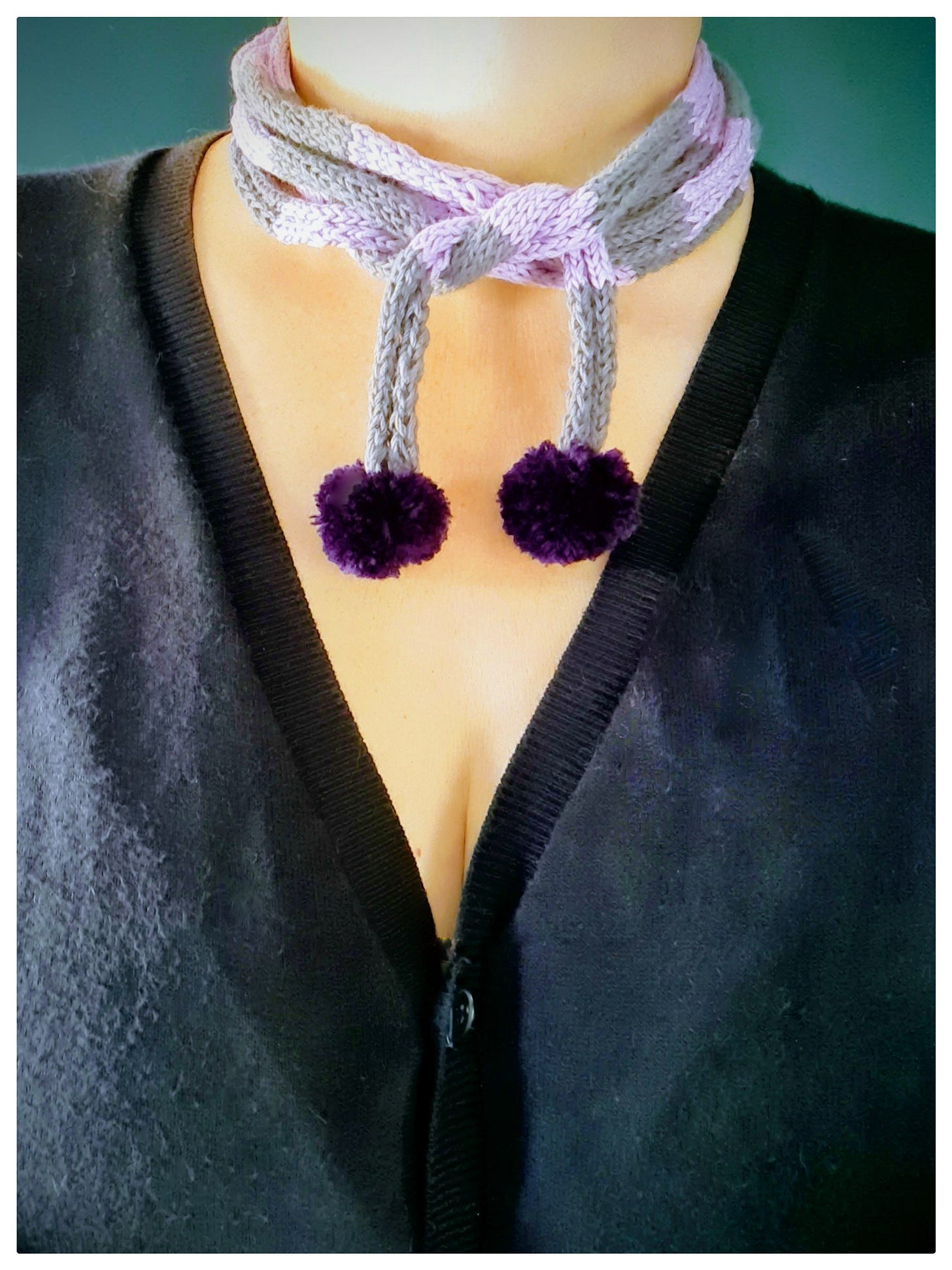 woman standing in front of a teal wall wears a hand knitted stripy neck scarf with pom poms and a navy v necked cardigan. The photo has been cropped at the neck.