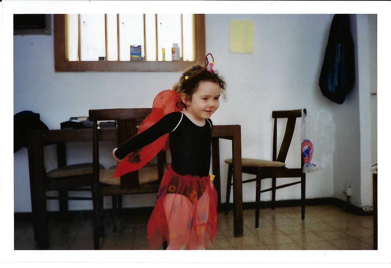 Picture of a little girl in a long-sleeved black leotard, with a tulle red skirt and red costume wings and red butterfly antennae, grinning for the camera
